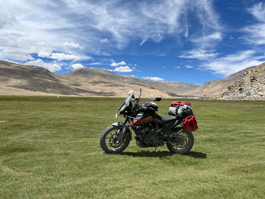 A motorcycle loaded with gear stands in a vast, green landscape with distant mountains under a bright blue sky.